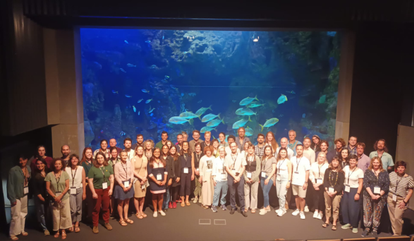 Group photo of 2023 Summer School in San Sebastian, Spain. The group is at the aquarium, standing in front of a tank of different fish