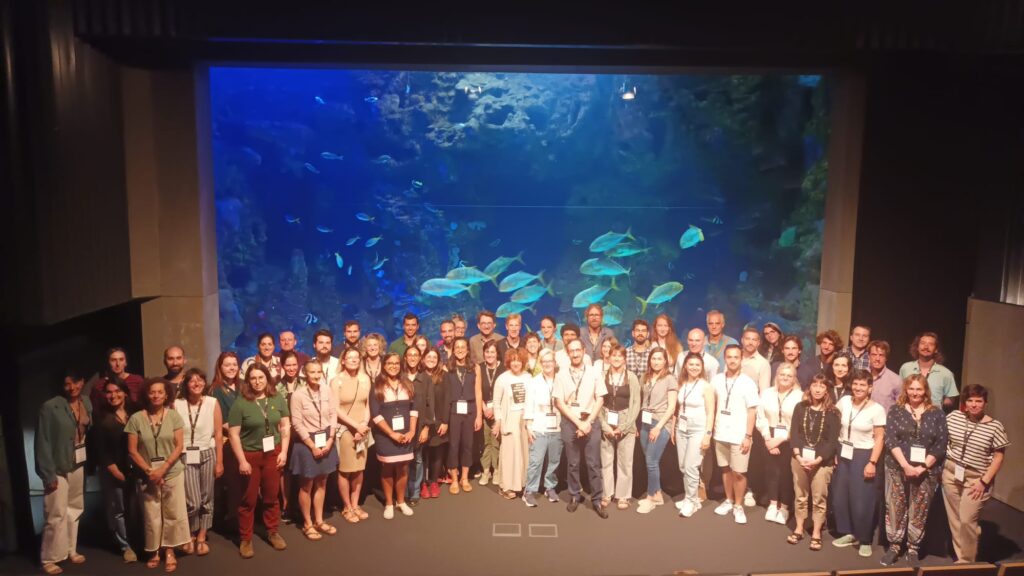Group photo of 2023 Summer School in San Sebastian, Spain. The group is at the aquarium, standing in front of a tank of different fish